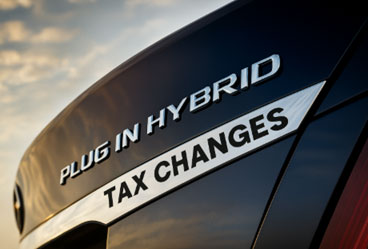 Close-up of a car badge reading Plug-in Hybrid with the words Tax Changes added below, symbolising upcoming taxation rule changes for hybrid vehicles against a cloudy sky background.